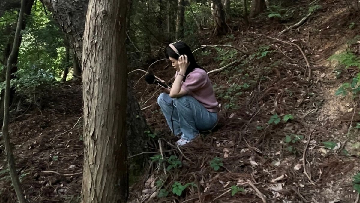 Hyun-Jin Yoo crouching in a forest wearing headphones and holding a recorder, field recording.
